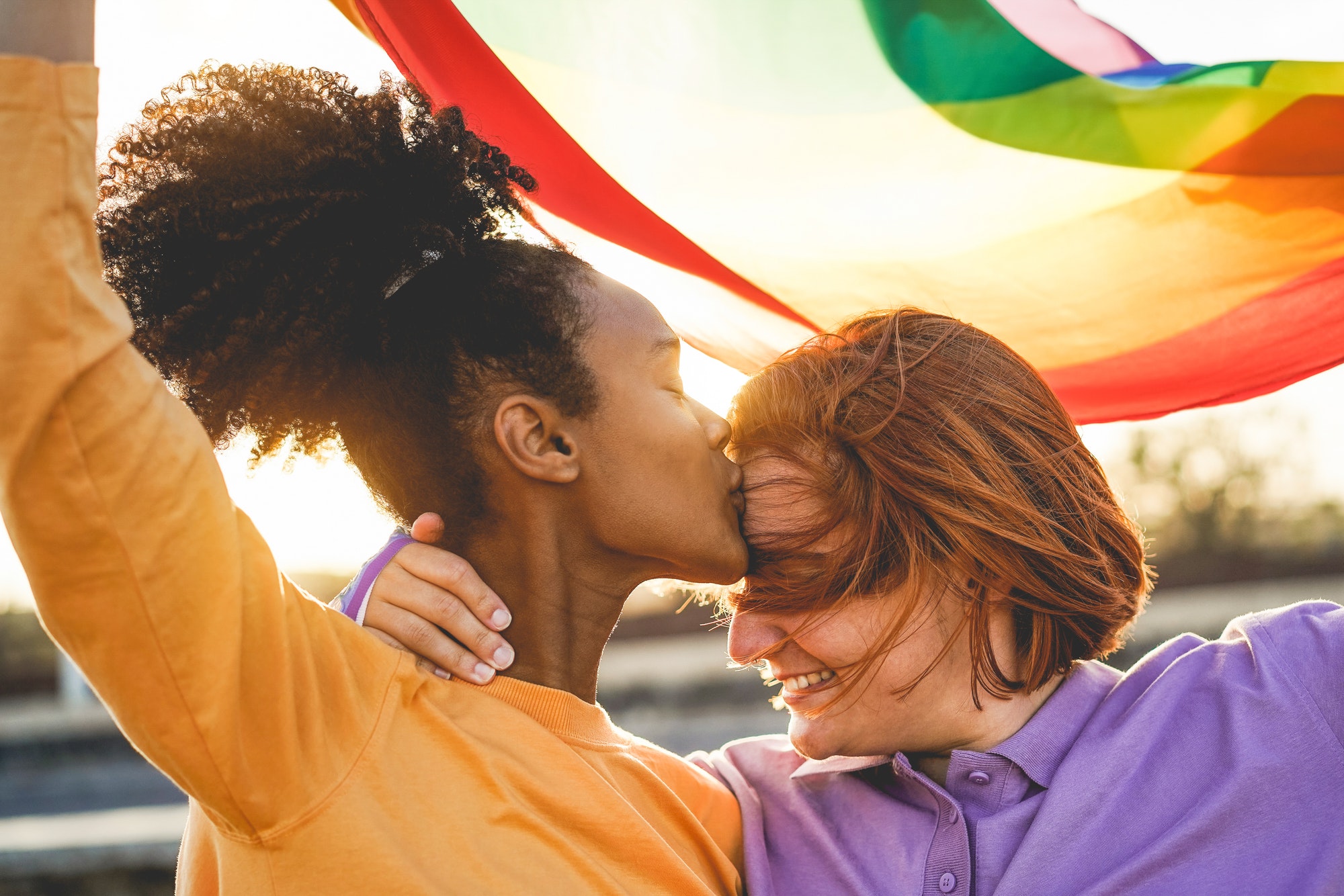 Happy women gay couple having tender moment holding rainbow flag outdoor